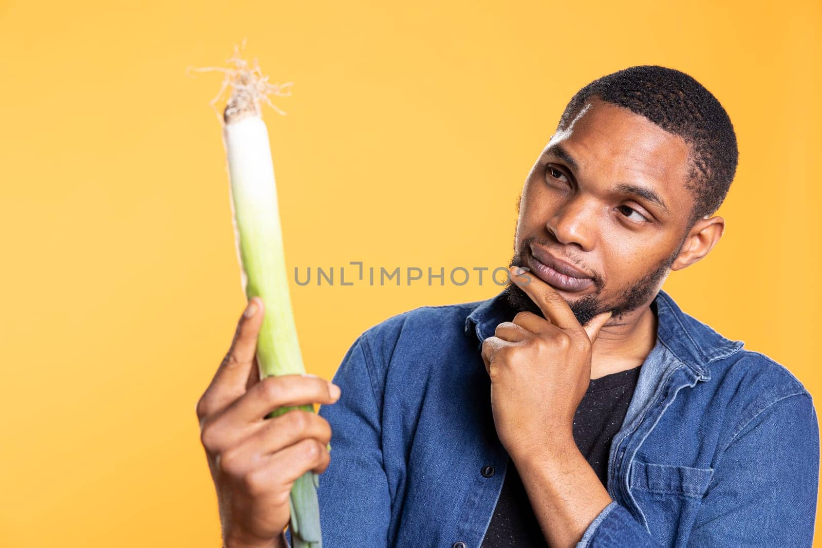 African american guy examines a locally grown leek plant against yellow background, doing a close inspection on green onion. Vegan person looking focused at organic food product.