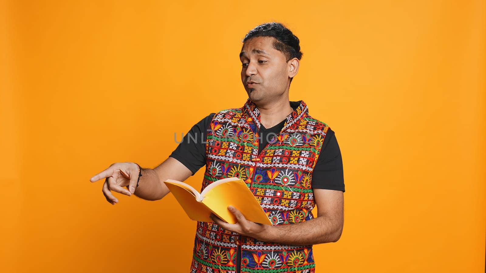 Joyous indian teacher reading book for pupils during class, isolated over studio background. Cheerful man holding novel, telling story to kindergarten kids, teaching them, camera B