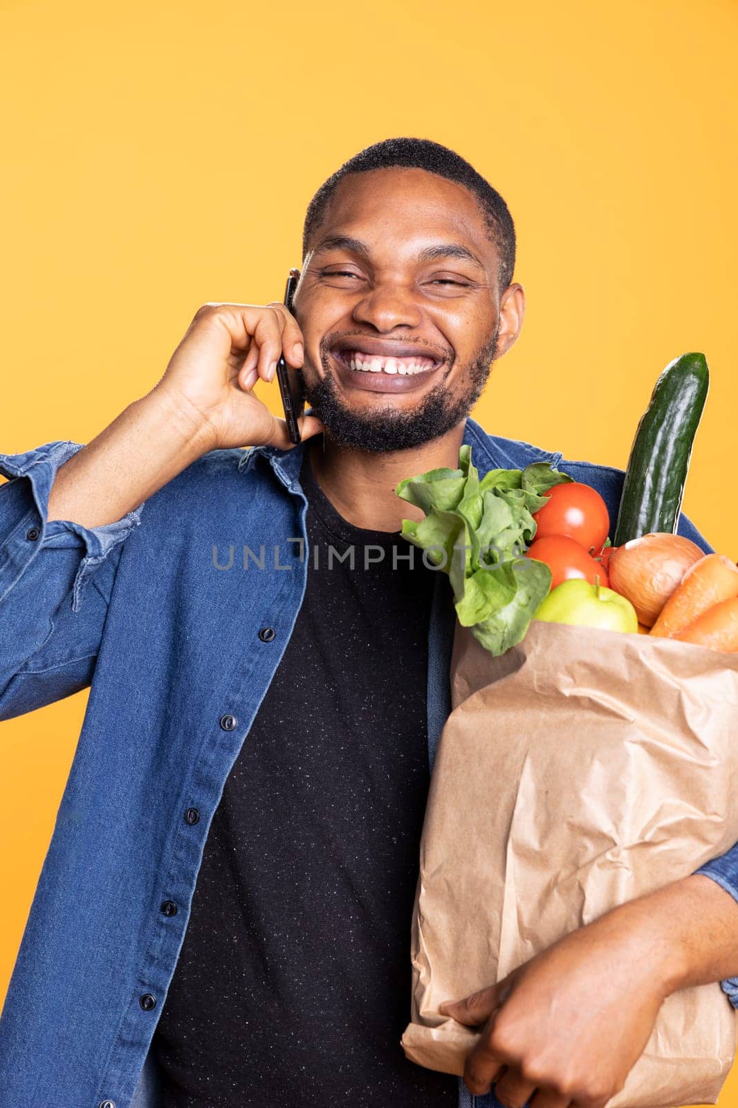 African american charismatic adult chats on his mobile device in studio, carries a paper bag full of groceries from local supermarket. Joyful friendly guy answers phone call during shopping.