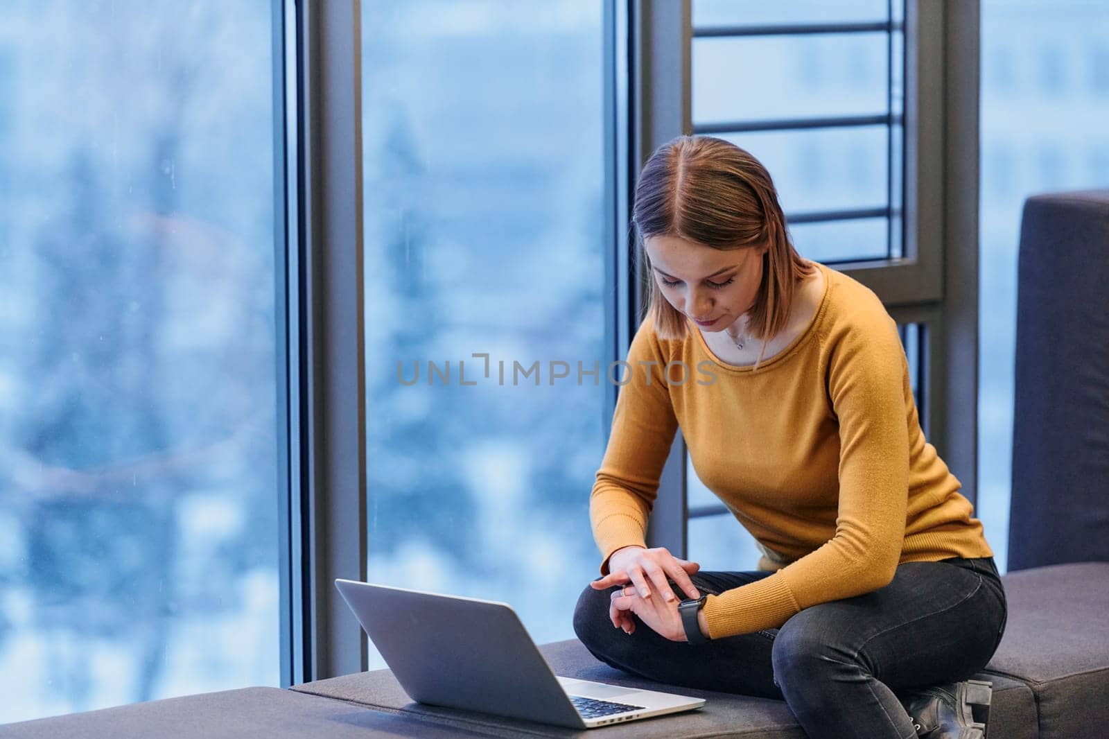 A businesswoman utilizes her laptop while seated by the window of a large corporate building, offering a picturesque view of the city skyline as her backdrop