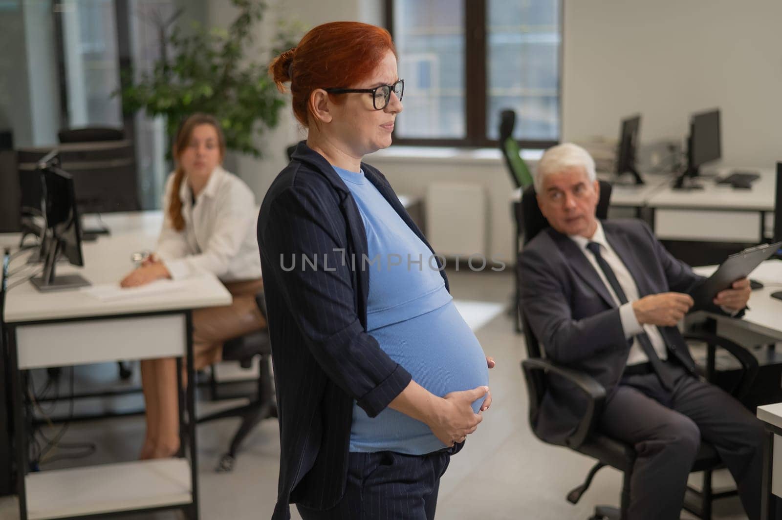 A pregnant woman suffers from pain and holds her stomach while standing in the middle of the office next to her colleagues. by mrwed54