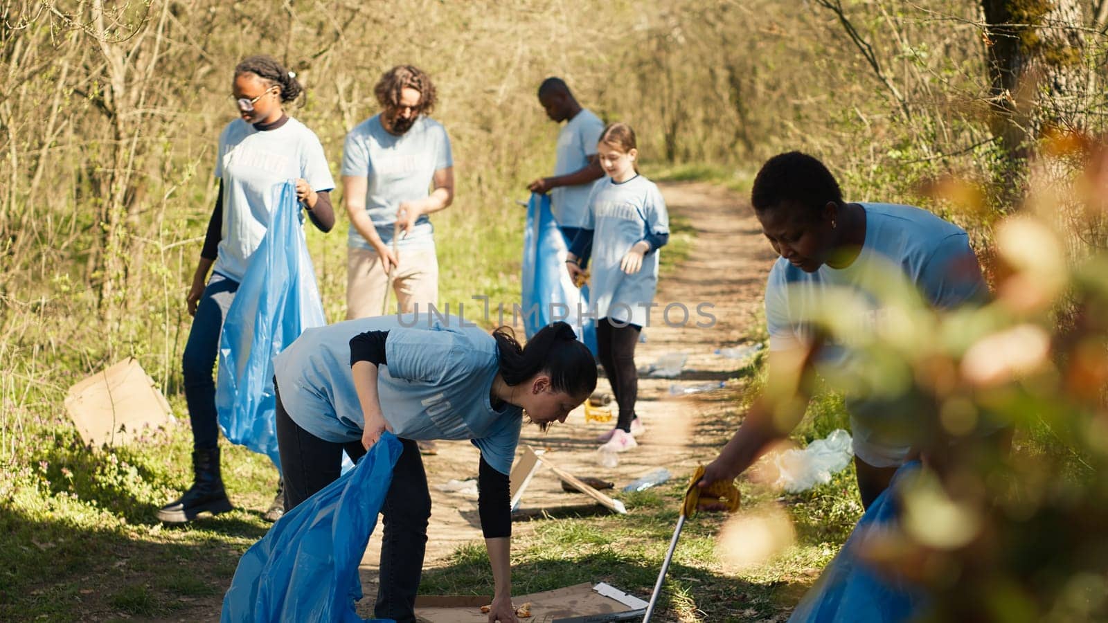 Diverse group of activists gathering to clean up a forest area, using litter cleanup tools like garbage bags and a long claw to grab the trash. People picking rubbish and recycling. Camera A.