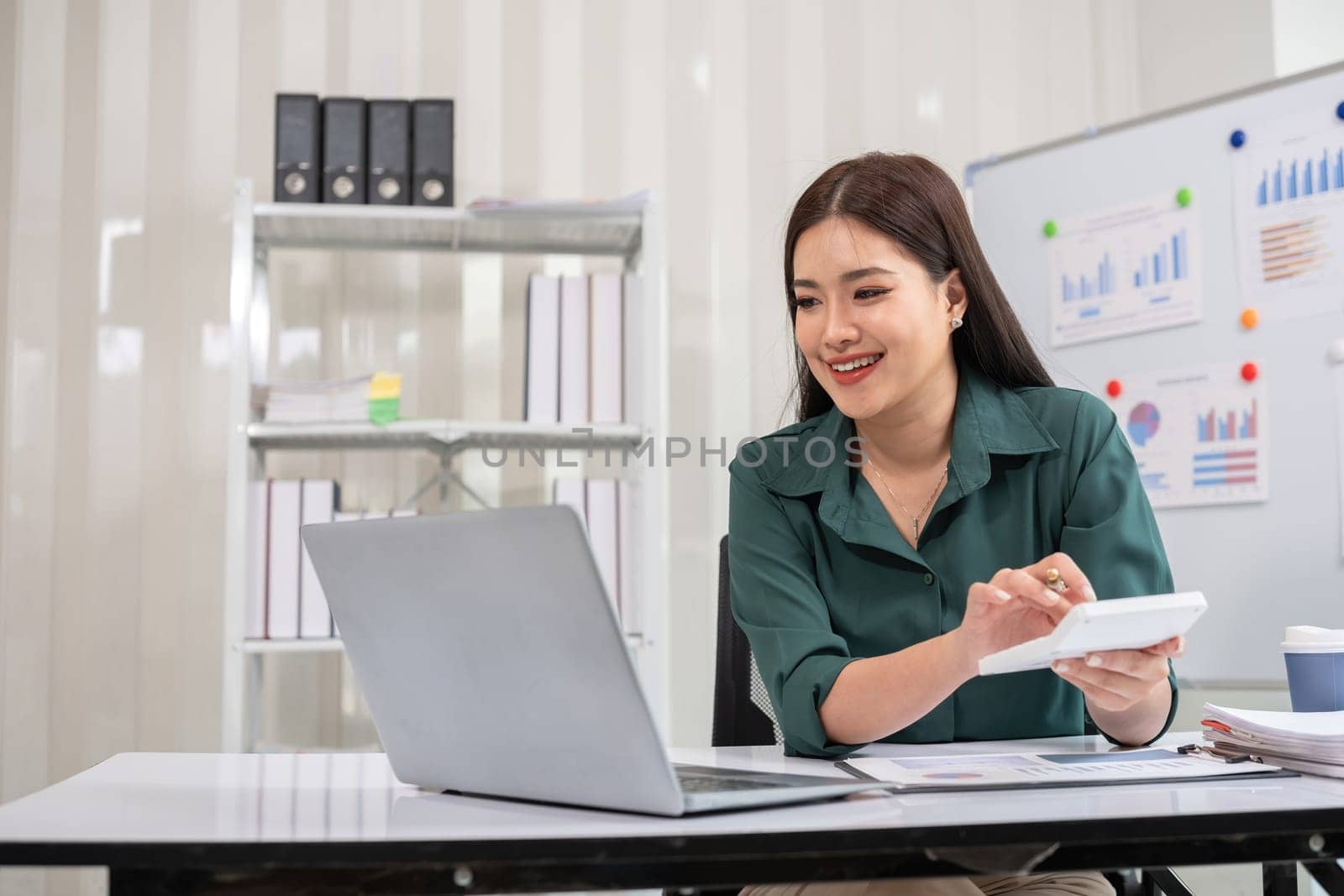 Asian businesswoman working on financial document with laptop on desk in office room.