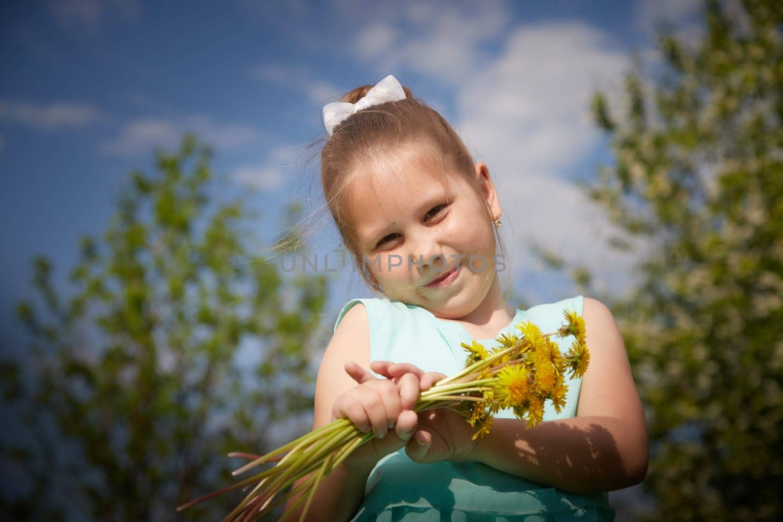 A little girl with a bouquet of yellow dandelion flowers and green trees with a blue sky in the background. Portrait of a happy Baby on a sunny summer day in nature by keleny