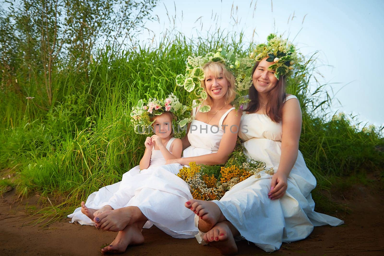 Ivan Kupala Celebration, photoshoot. Portrait of Girls With Floral Wreaths by the River at Sunset. Family clad in white dresses celebrate at dusk by keleny
