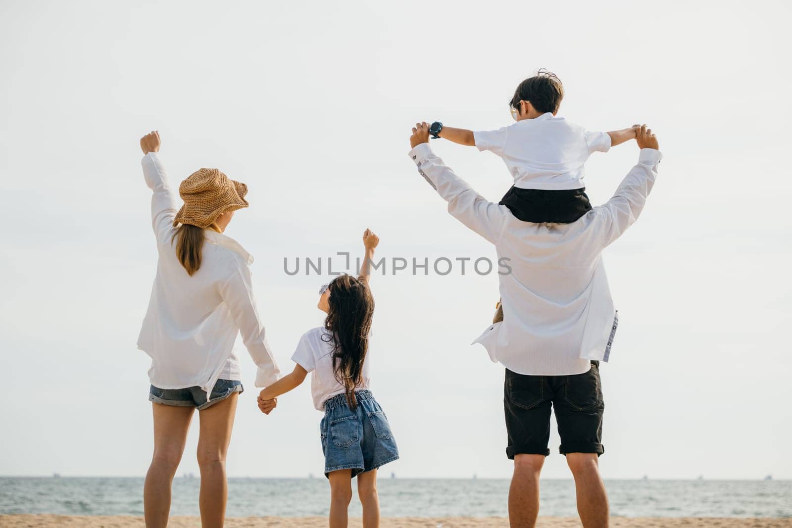 Amidst sunset glow a family of four stands joyfully on the sea beach. Father carries his son on shoulders creating heartwarming image of togetherness and carefree beach fun. Family on beach vacation