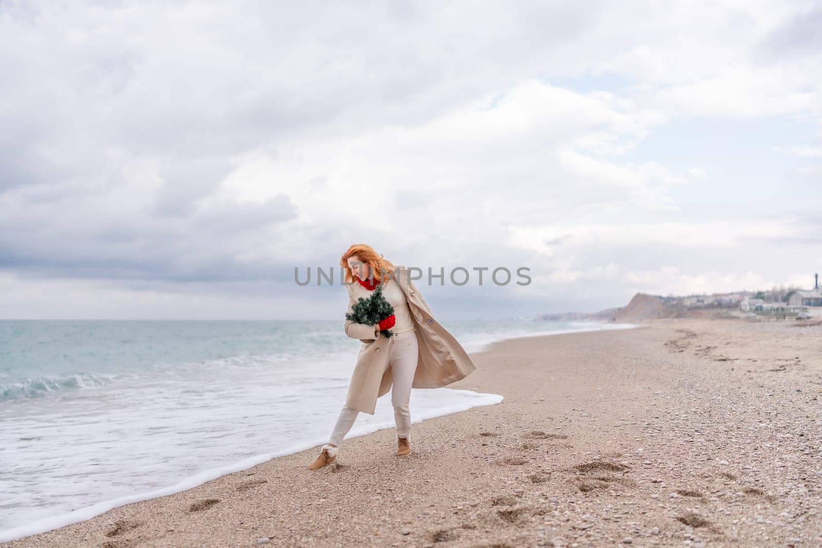 Redhead woman Christmas tree sea. Christmas portrait of a happy redhead woman walking along the beach and holding a Christmas tree in her hands. Dressed in a light coat, white suit and red mittens. by Matiunina