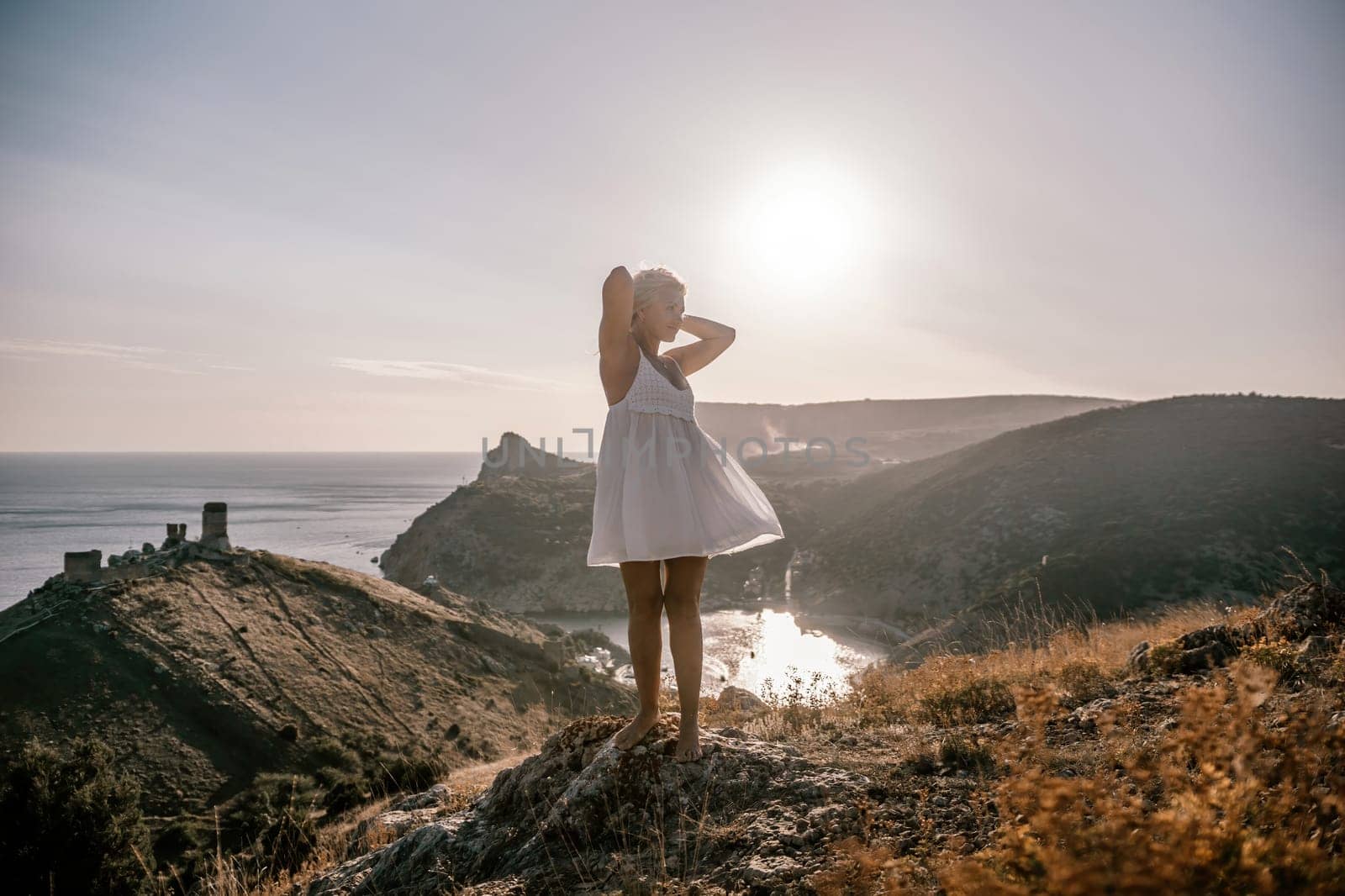A blonde woman stands on a hill overlooking the ocean. She is wearing a white dress and she is enjoying the view