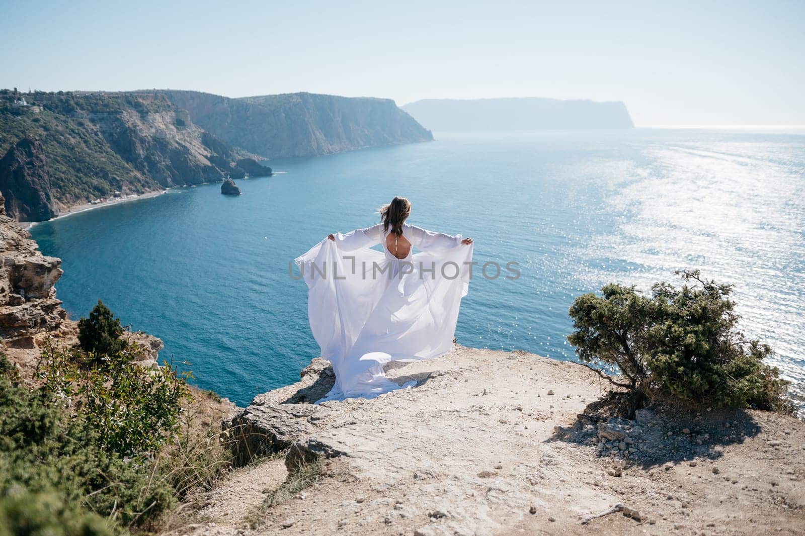 woman white dress stands on a rocky cliff overlooking the ocean. The scene is serene and peaceful, with the woman's dress billowing in the wind. The ocean in the background is calm and blue. by Matiunina