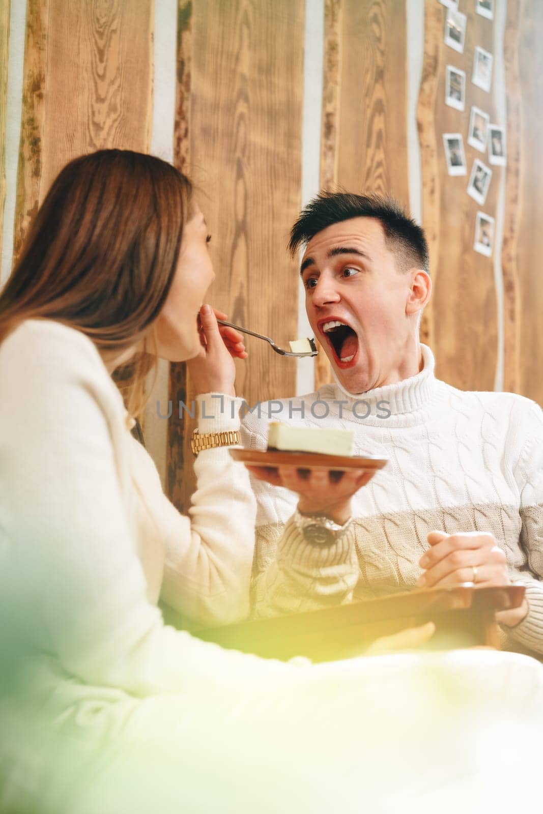 A joyful young man with an open mouth is eagerly anticipating a taste of dessert from a spoon held by his companion. They are seated in a warm and rustic wooden cabin, surrounded by a cozy ambiance and the soft glow of natural light. The moment captures casual domestic bliss and the simple pleasure of sharing food.