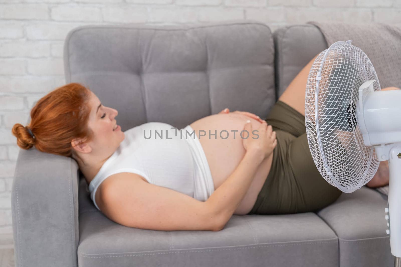 A pregnant woman is lying on the couch, stroking her tummy and enjoying the cool air from the electric fan. by mrwed54