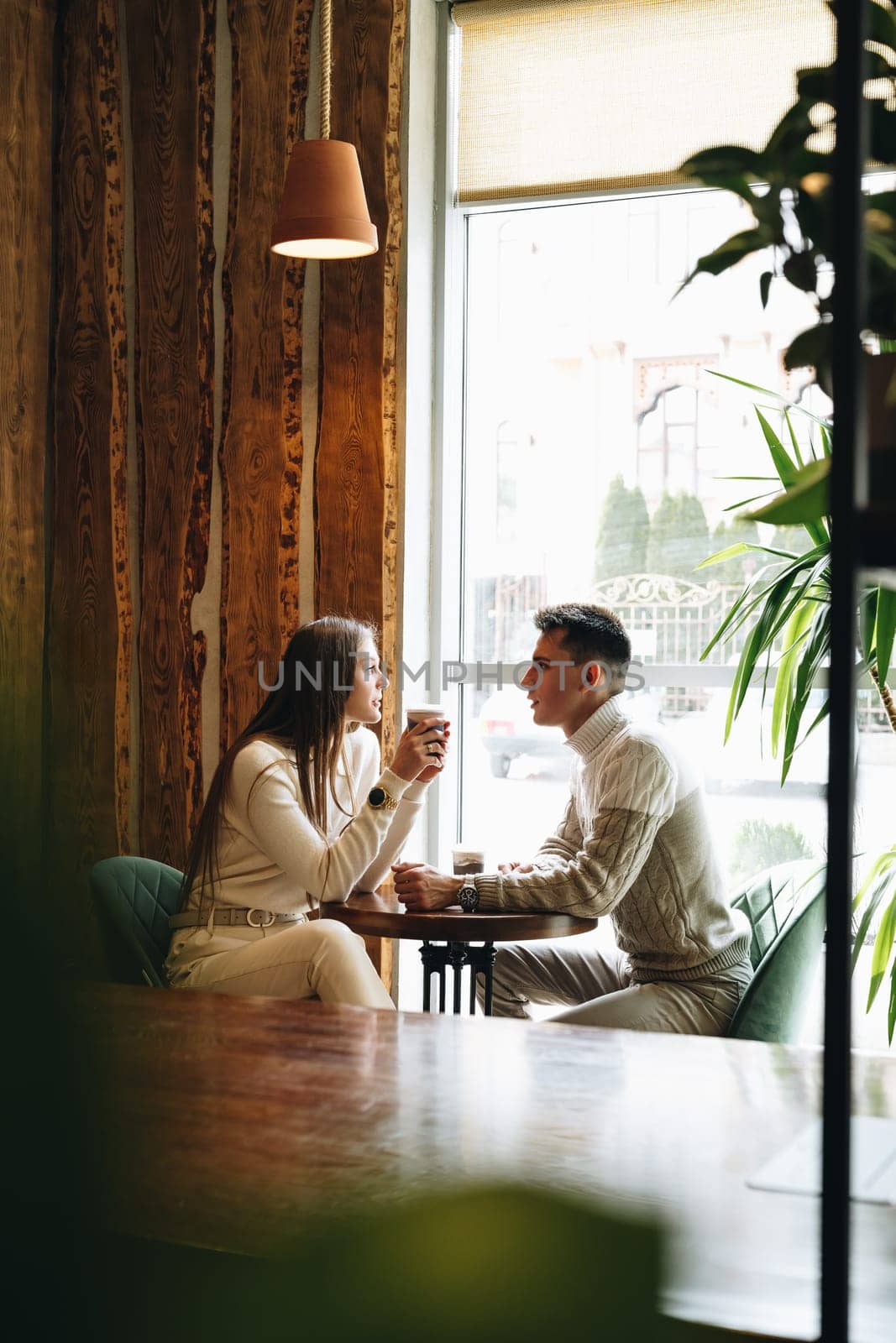 Two professionals are engaged in a conversation while sitting opposite each other at a wooden table in a cafe with natural light streaming in from a large window. They appear relaxed and focused, with cups of coffee in hand, surrounded by a warm, rustic interior.