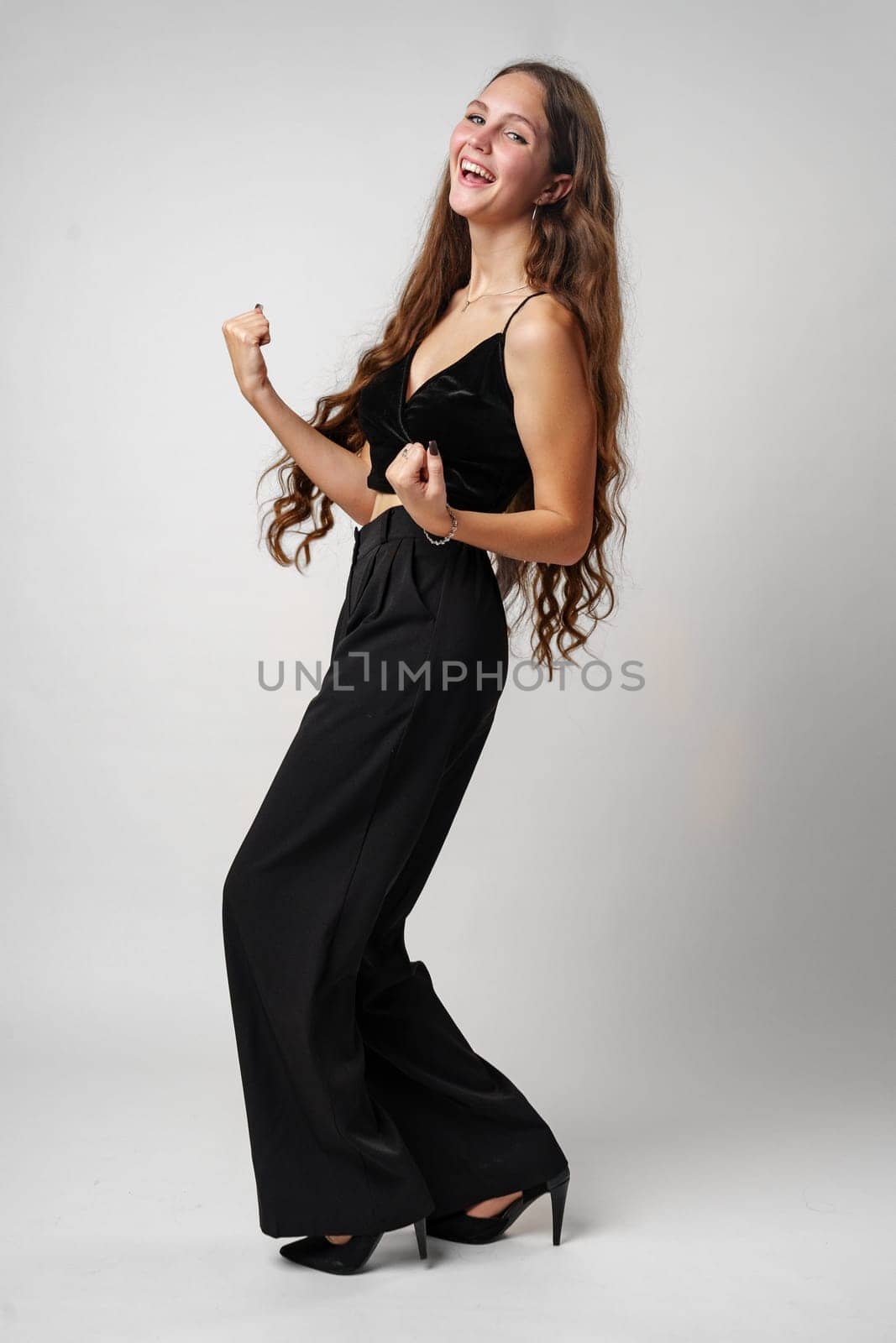 A radiant young woman with long hair is captured in a moment of celebration, wearing an elegant black dress with a sleeveless top and full-length skirt. She appears to be dancing or celebrating, with an ecstatic expression on her face and her hands raised in a gesture of excitement. The background is simple and neutral, highlighting the subjects joyous demeanor and stylish attire.