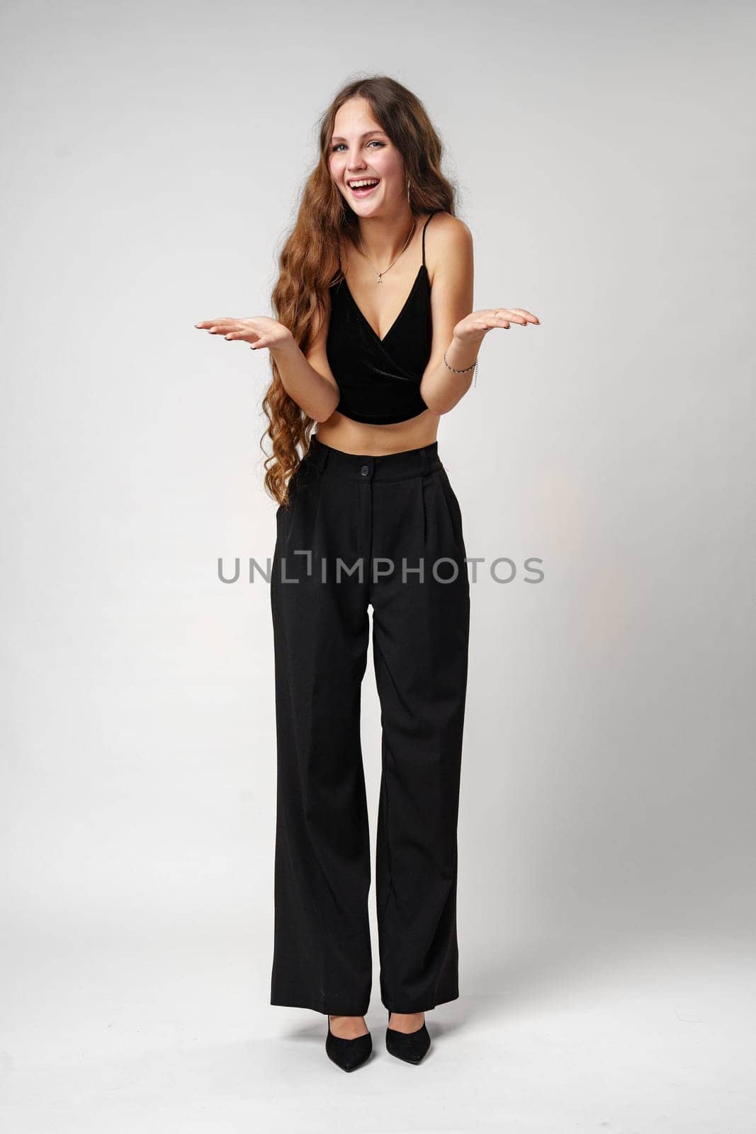 A young woman with long, wavy hair smiles broadly and gestures animatedly with her hands, expressing excitement and happiness. She is wearing a black V-neck top and stands against a simple, uncluttered backdrop that accentuates her lively demeanor.