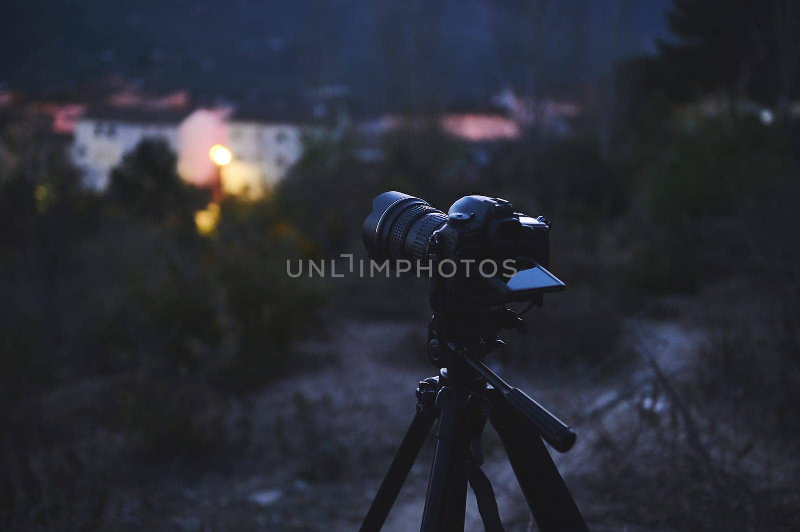 A digital camera with black mockup touch screen placed on tripod by a travel photographer hiker, capturing the beautiful view of a city in mountains at sunrise. The concept of World photography Day