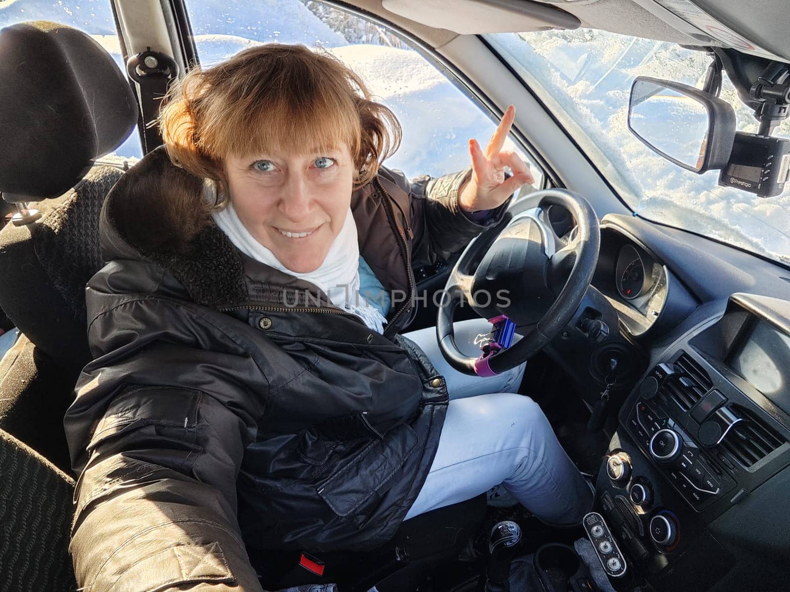 A woman in a car taking a selfie while enjoying a drive. Female driver in warm jacket posing inside car in warm jacket on cold day by keleny