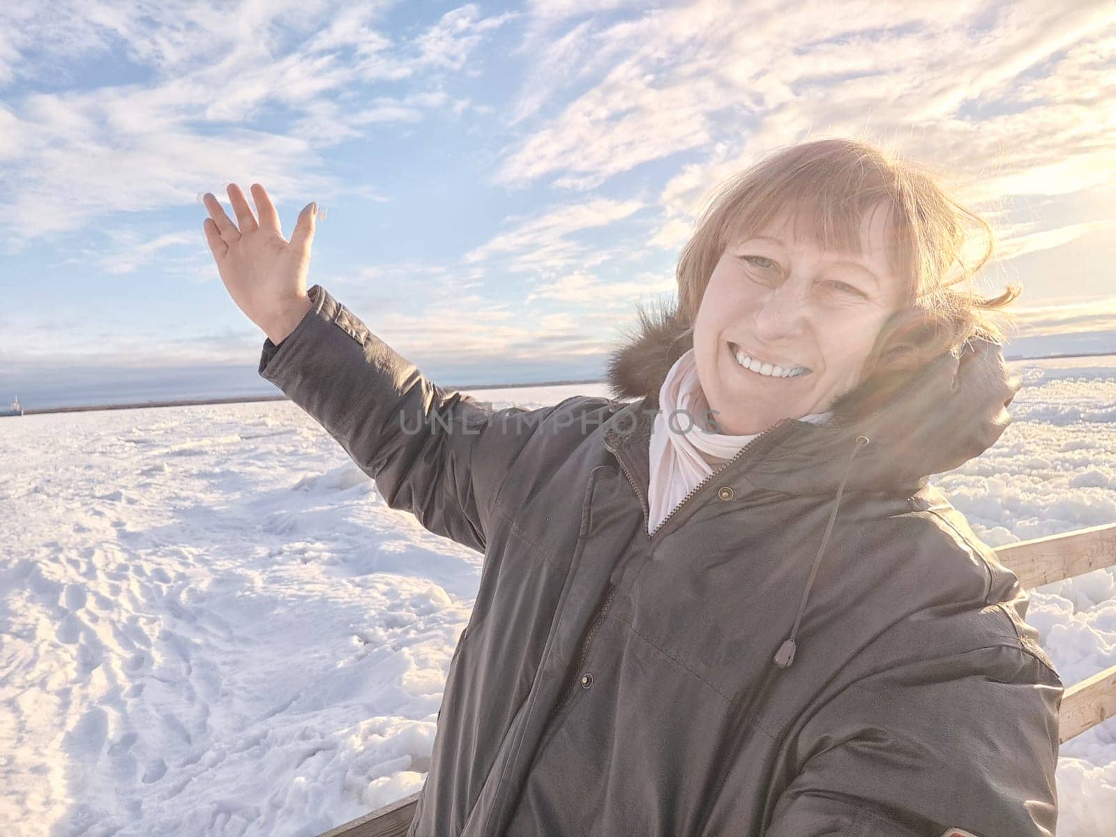 A cheerful middle aged woman in a winter coat taking selfie on nature outdoors in sunny day with blue sky