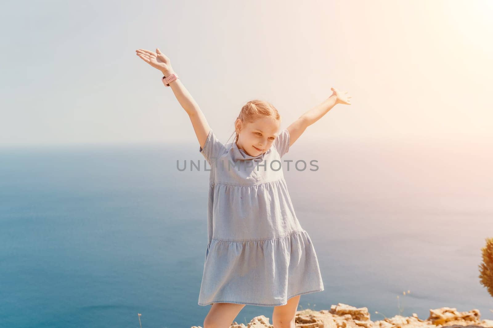 A young girl is standing on a beach, wearing a blue dress and holding her arms up in the air. Concept of joy and freedom, as the girl appears to be enjoying her time by the ocean. by panophotograph