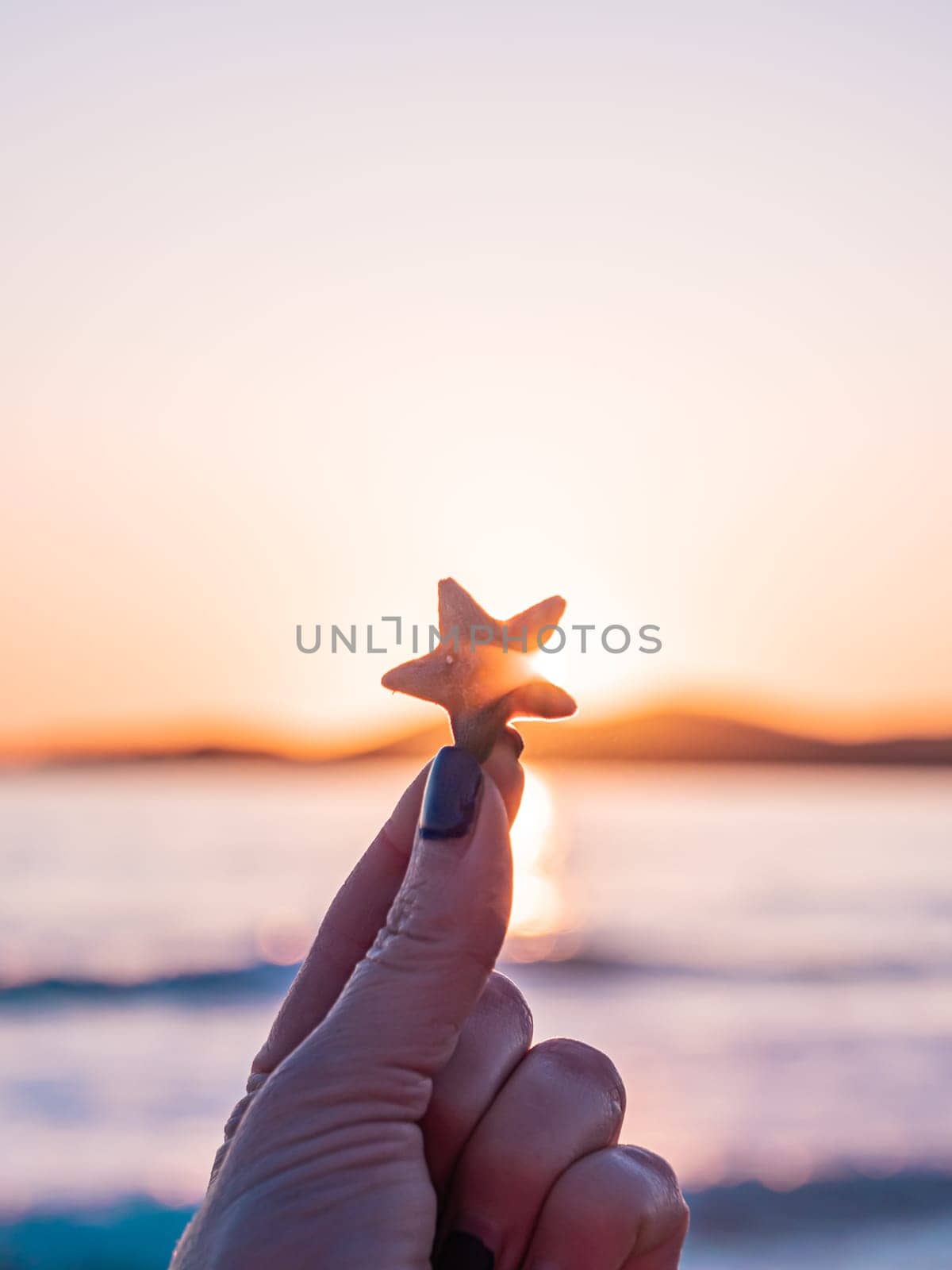 Hand holding small starfish against colorful beach sunset horizon by Busker