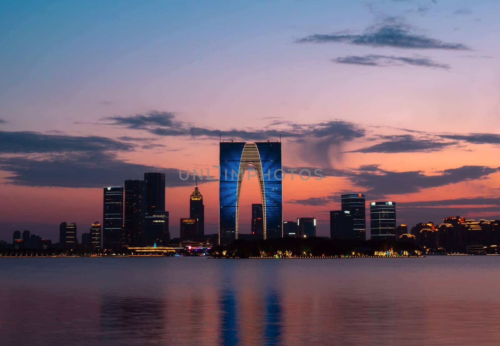 The Gate of the Orient, a distinctive skyscraper in Suzhou, China, is illuminated against the evening sky as it overlooks a calm lake. Surrounding buildings and city lights reflect on the water, creating a picturesque urban scene.