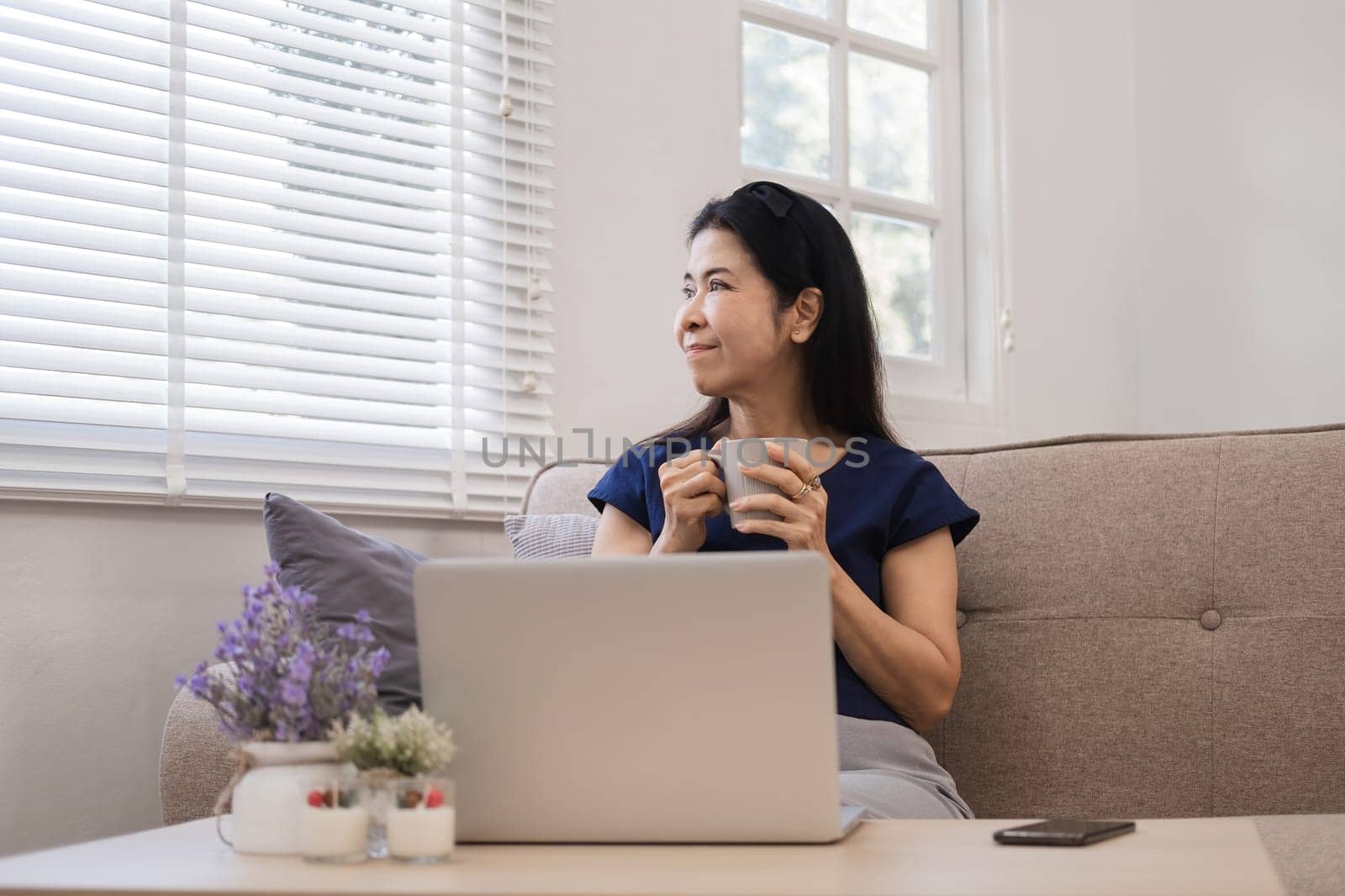 Happy Asian retired woman in her 60's enjoying social media on laptop while relaxing in living room. by wichayada