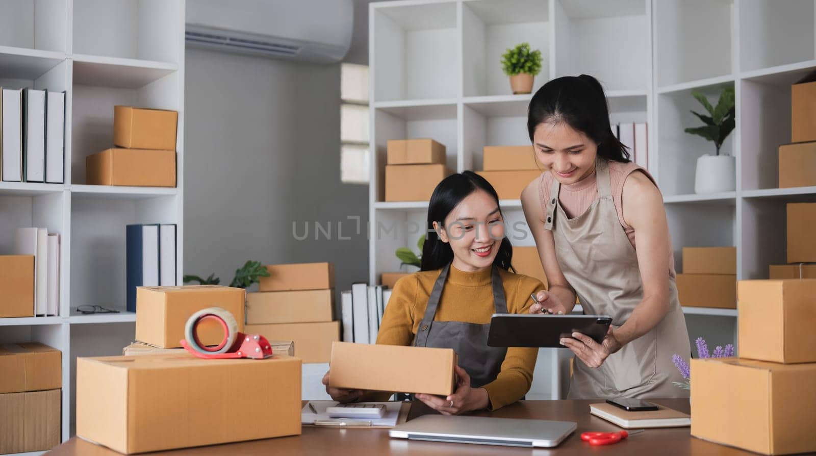Two Asian women packing boxes and using a tablet in a home office. Concept of small business and e-commerce..