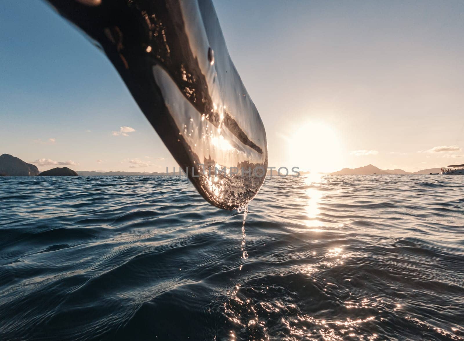 A kayak paddle drips water as the sun sets over a peaceful ocean, creating a tranquil and picturesque scene. The horizon is adorned with distant mountains, reflecting the golden hue of the setting sun.