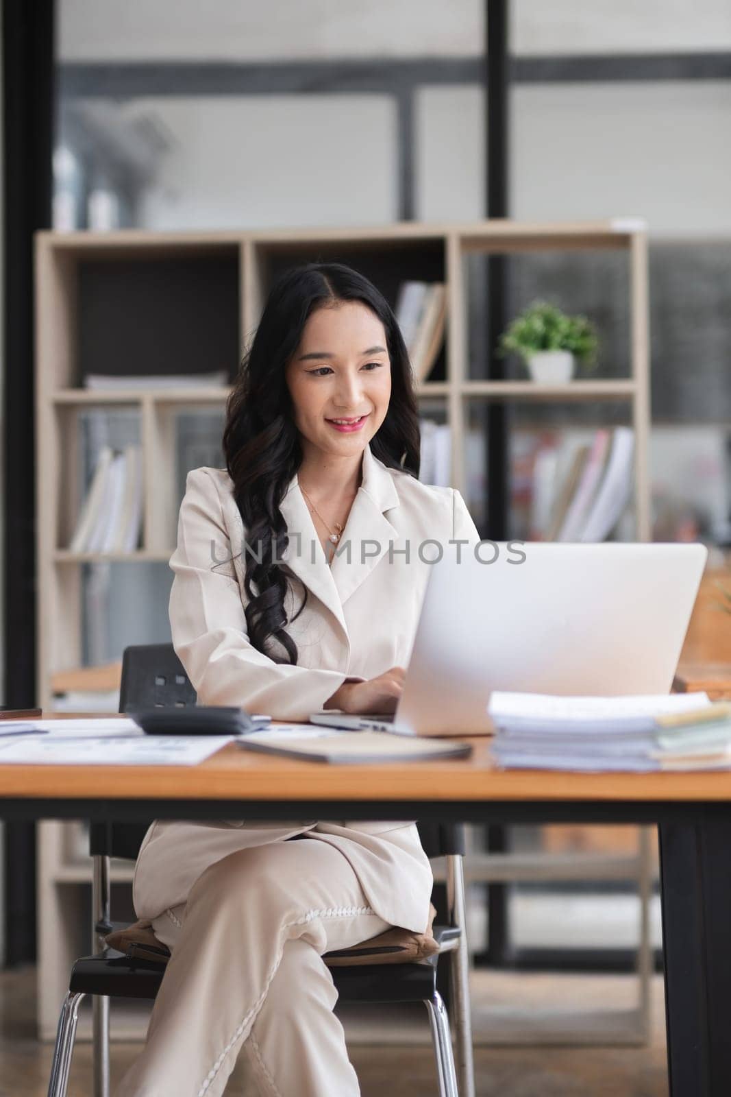 Asian businesswoman working on financial document with laptop on desk in office room.