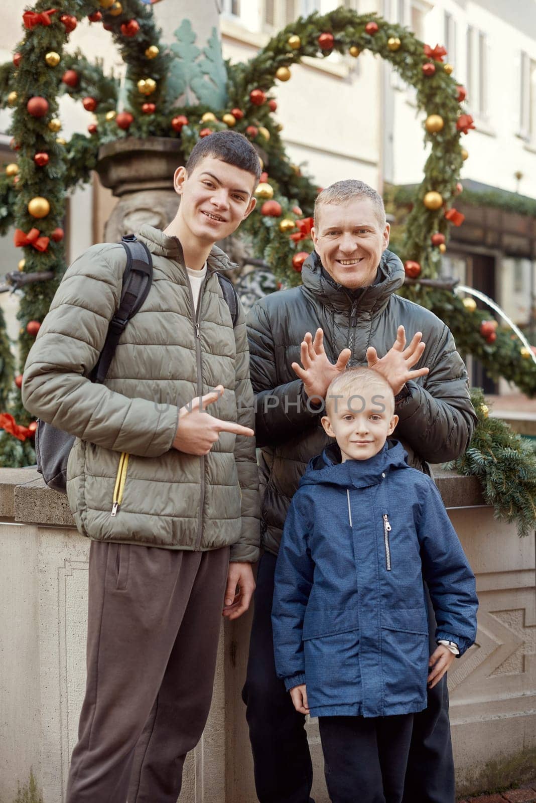 Joyful Family Portrait: Father and Two Sons by Festive Vintage Fountain. Capture the essence of familial happiness with this heartwarming image featuring a handsome father with his two sons standing against the backdrop of a festively decorated vintage fountain. The image beautifully blends the joy of the holiday season with the timeless charm of family togetherness. The father is 44 years old, the elder son is 17, and the younger one is 8, creating a memorable and multigenerational festive scene.