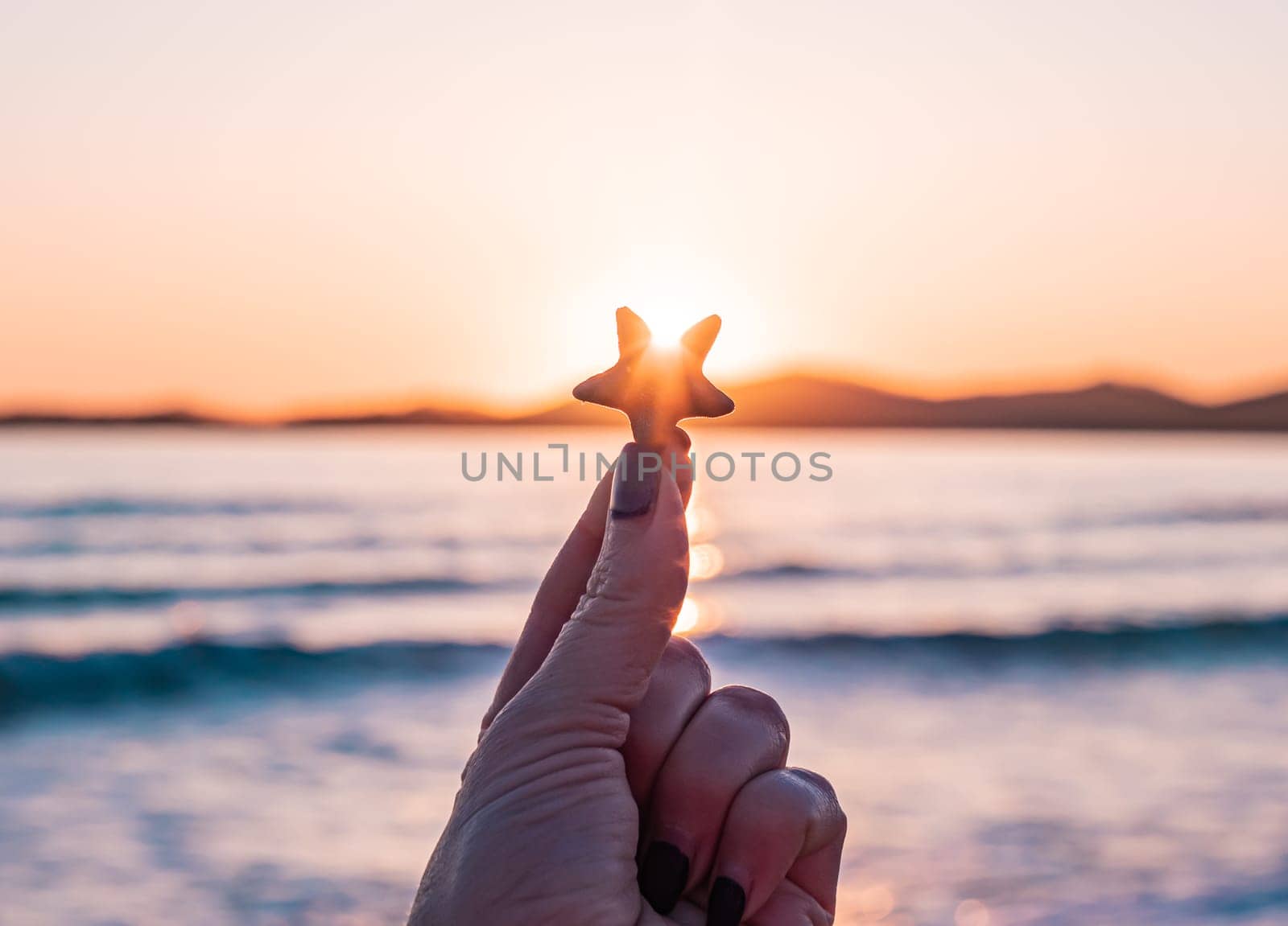 A hand with dark nail polish holds a starfish up towards the sunset, with the ocean and mountains in the background. The sky is filled with vibrant colors as the sun sets over the water.