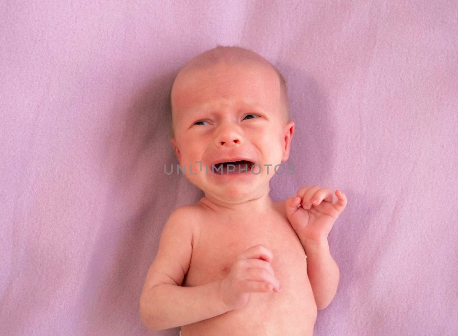 Newborn baby crying on a soft pink blanket in the afternoon light by Busker