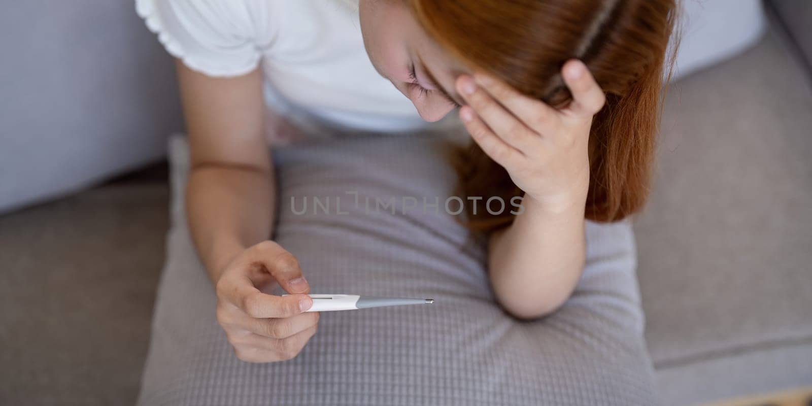 Young Asian woman with fever checking her temperature with a thermometer at home. Concept of illness and healthcare by nateemee