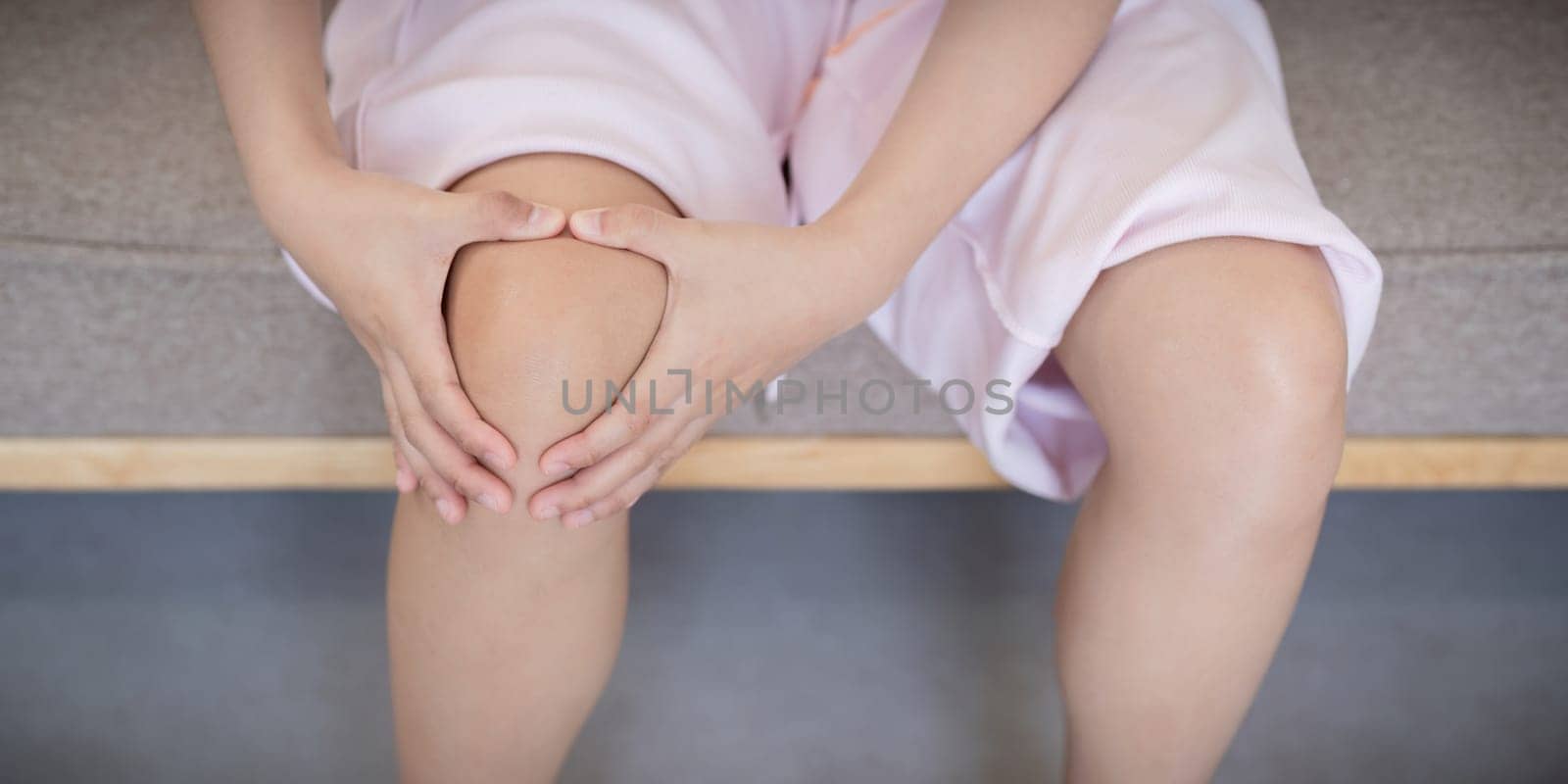 Close up of young Asian woman holding her knee in pain while sitting on a sofa. Concept of knee pain and health issues.