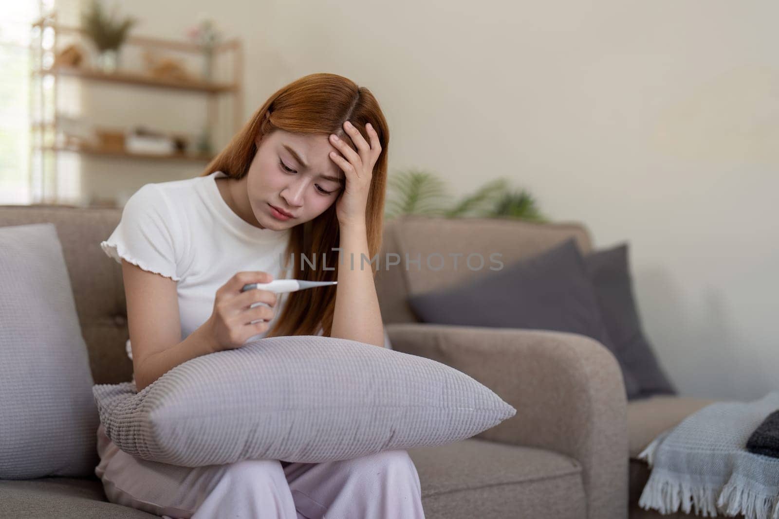 Young Asian woman with fever checking her temperature with a thermometer at home. Concept of illness and healthcare.