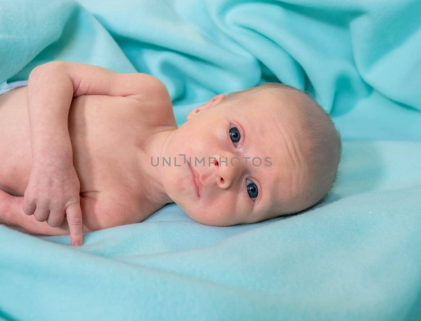 A newborn baby lies comfortably on a soft, aqua-colored blanket in a hospital room during the day. The baby gazes curiously at the camera, appearing calm and peaceful.