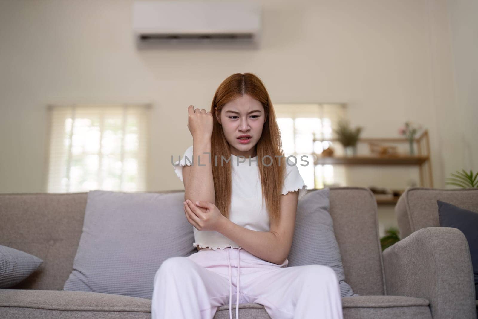 Young Asian woman experiencing arm pain while sitting on a sofa at home. Concept of health issues and discomfort.