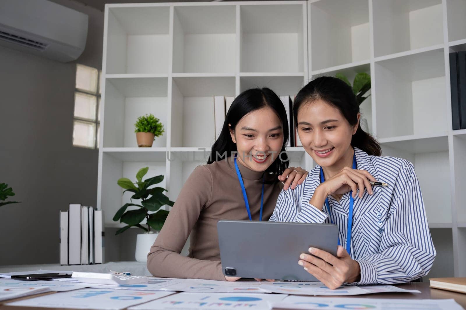 Two Asian businesswomen discussing work using a tablet in a modern office. Concept of teamwork and professional collaboration by wichayada