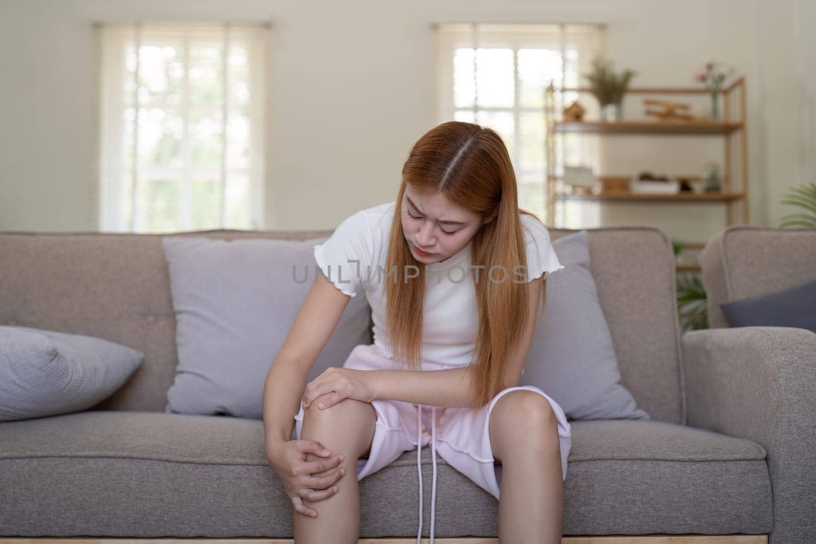 Young Asian woman experiencing knee pain while sitting on a sofa at home. Concept of health issues and discomfort.