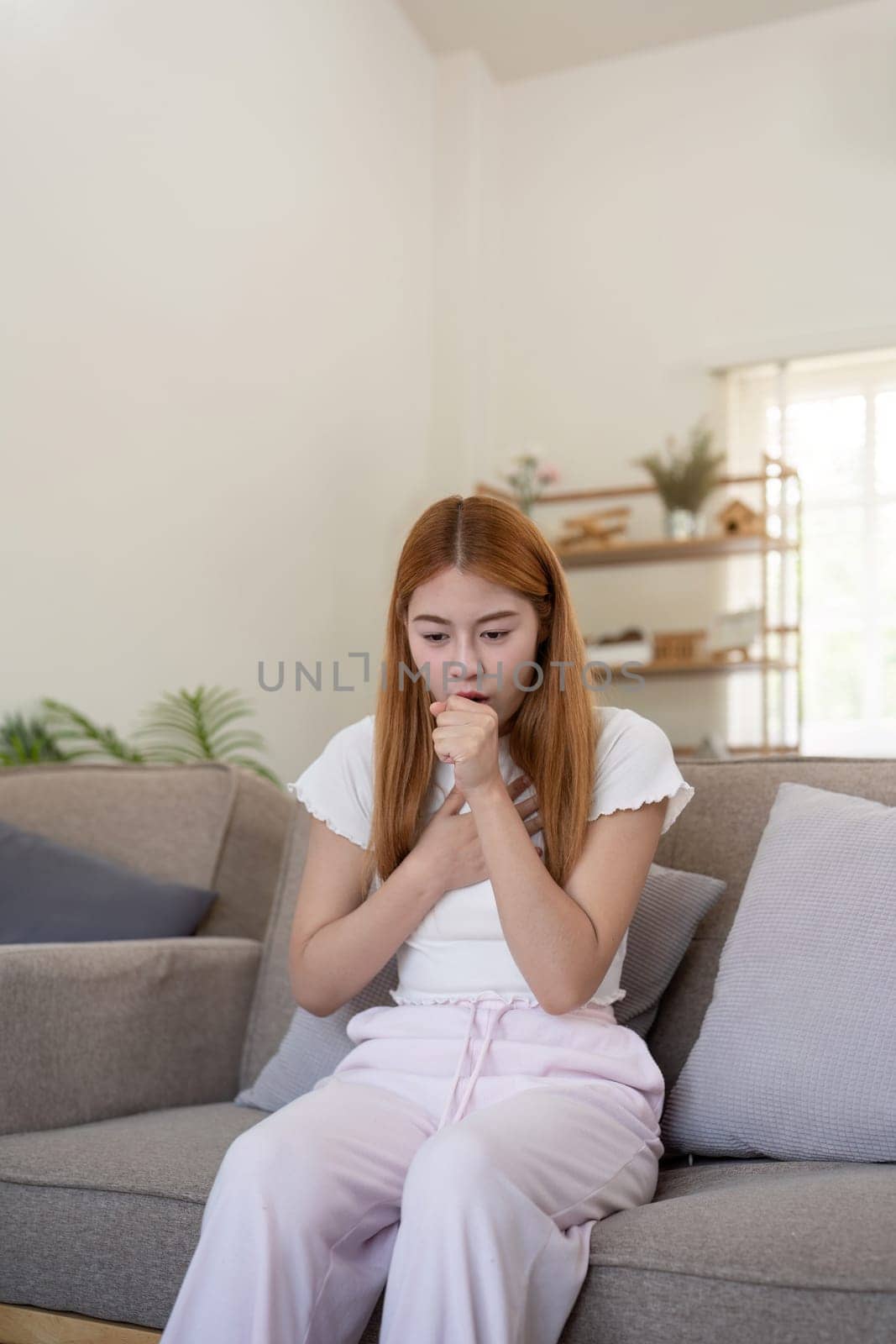 Young Asian woman coughing and holding her chest while sitting on a sofa. Concept of respiratory illness and health issues by nateemee