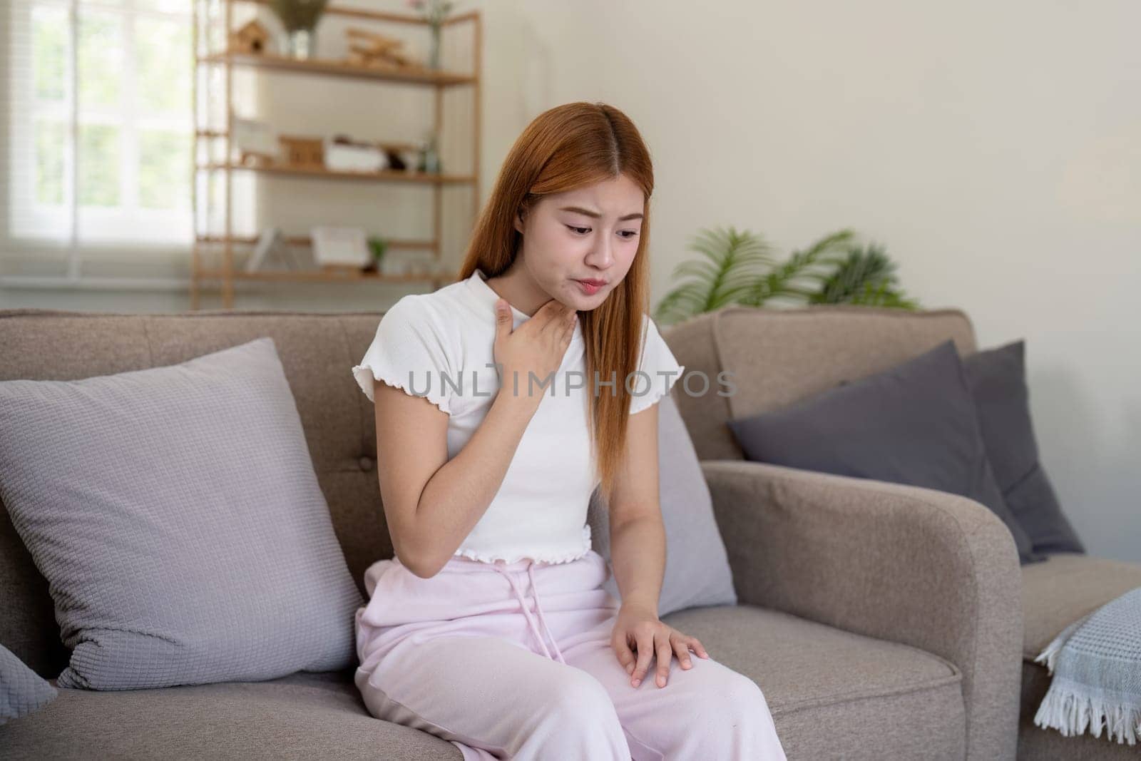 Young Asian woman holding her throat while sitting on a sofa. Concept of throat pain and health issues.