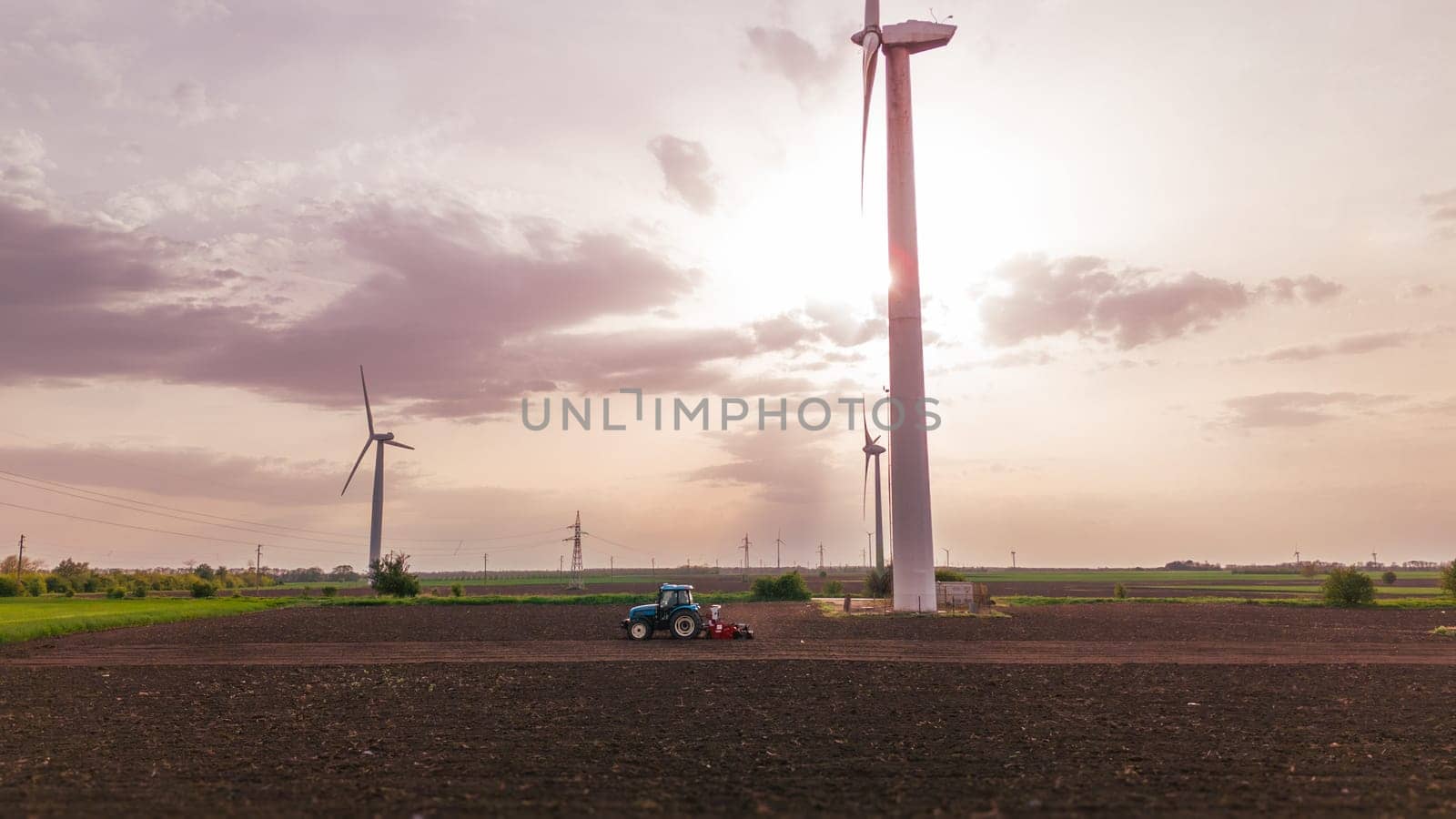 Seeds to sow. A tractor sows seeds against a background of meadows with wind turbines at sunset