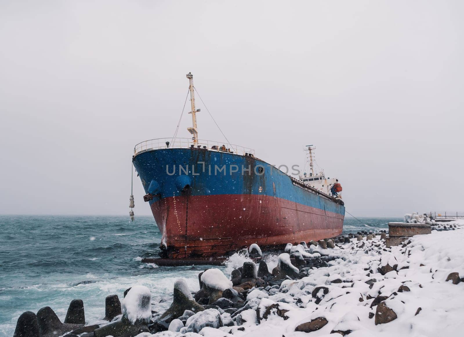 Cargo ship strands aground on a rocky shore after a storm, surrounded by snow and heavy waves. by Busker