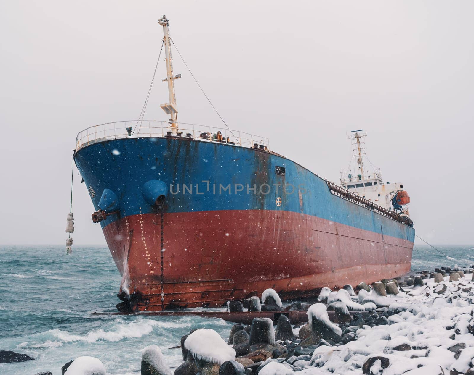 Cargo ship strands aground on a rocky shore after a storm, surrounded by snow and heavy waves. by Busker