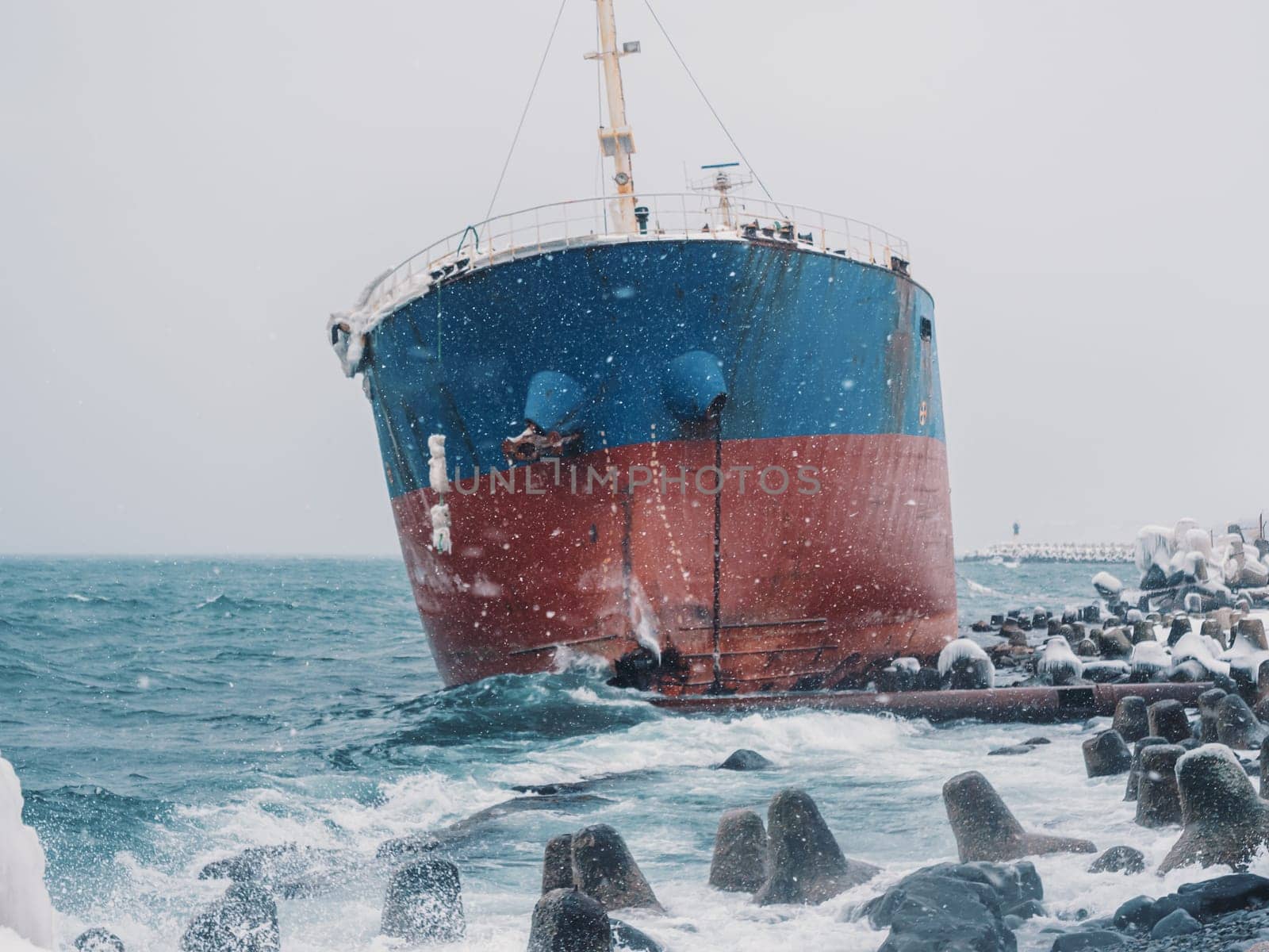 Cargo ship strands aground on a rocky shore after a storm, surrounded by snow and heavy waves.