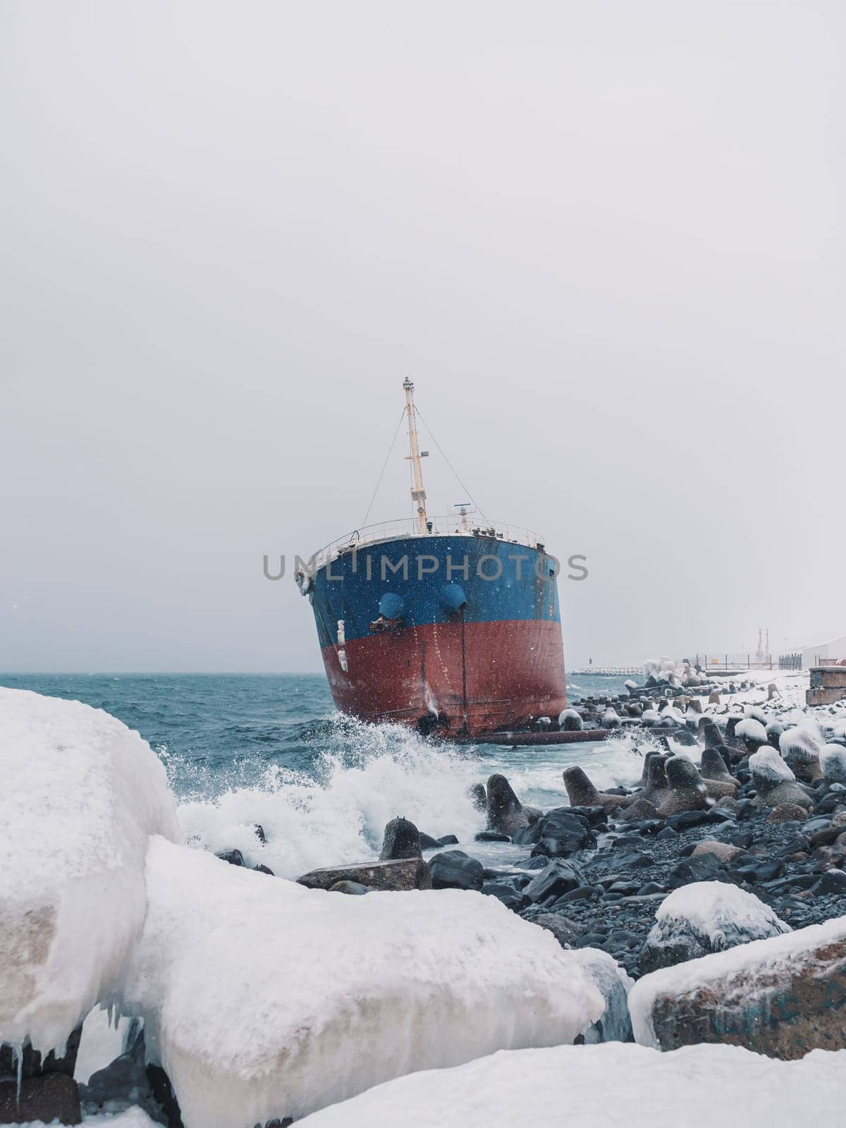 Cargo ship strands aground on a rocky shore after a storm, surrounded by snow and heavy waves.