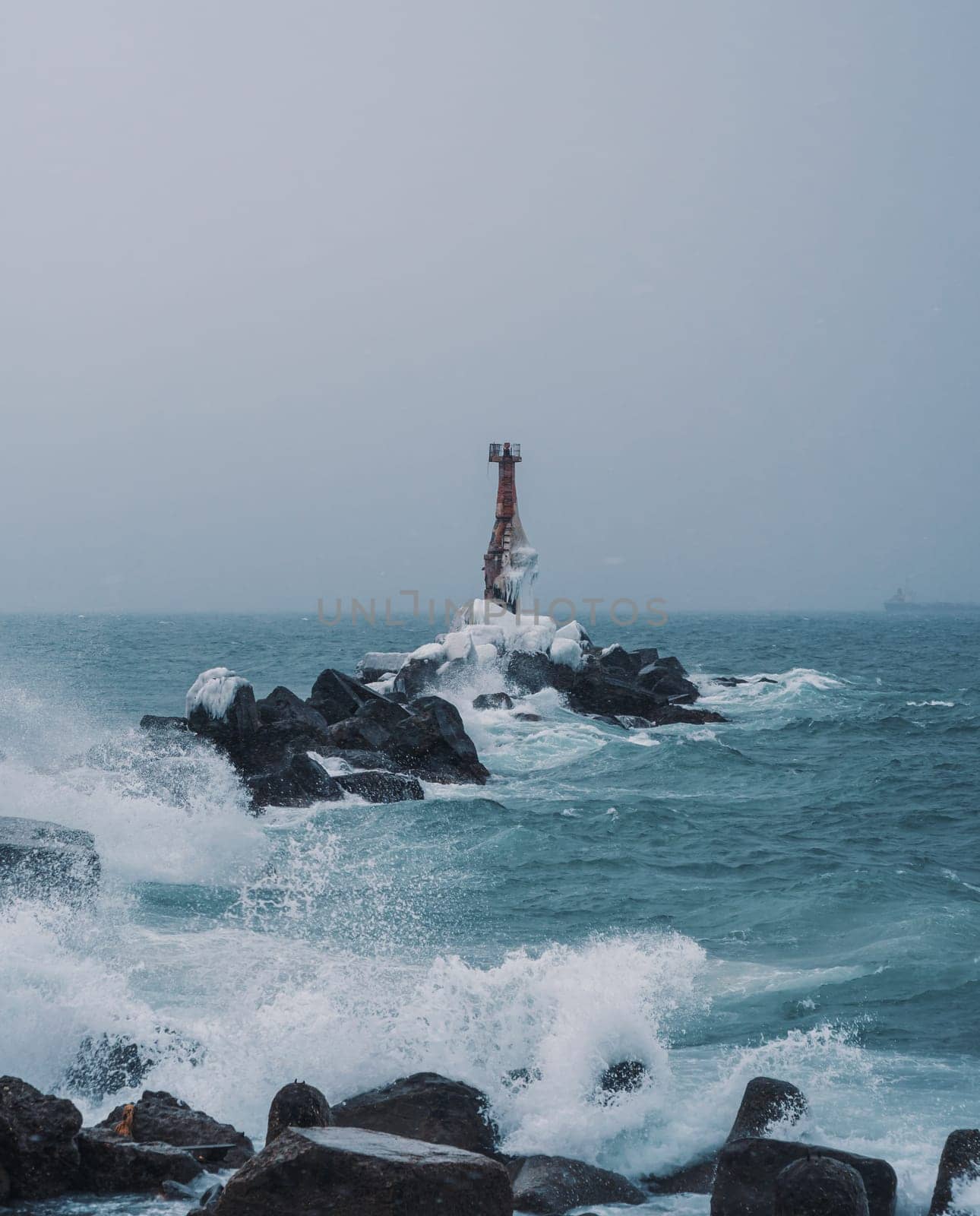 A solitary lighthouse stands firm on a rocky shore as waves crash against the rocks. The overcast sky and turbulent sea create a dramatic and moody atmosphere..