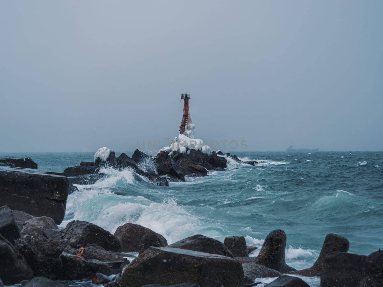 A solitary lighthouse stands firm on a rocky shore as waves crash against the rocks. The overcast sky and turbulent sea create a dramatic and moody atmosphere..