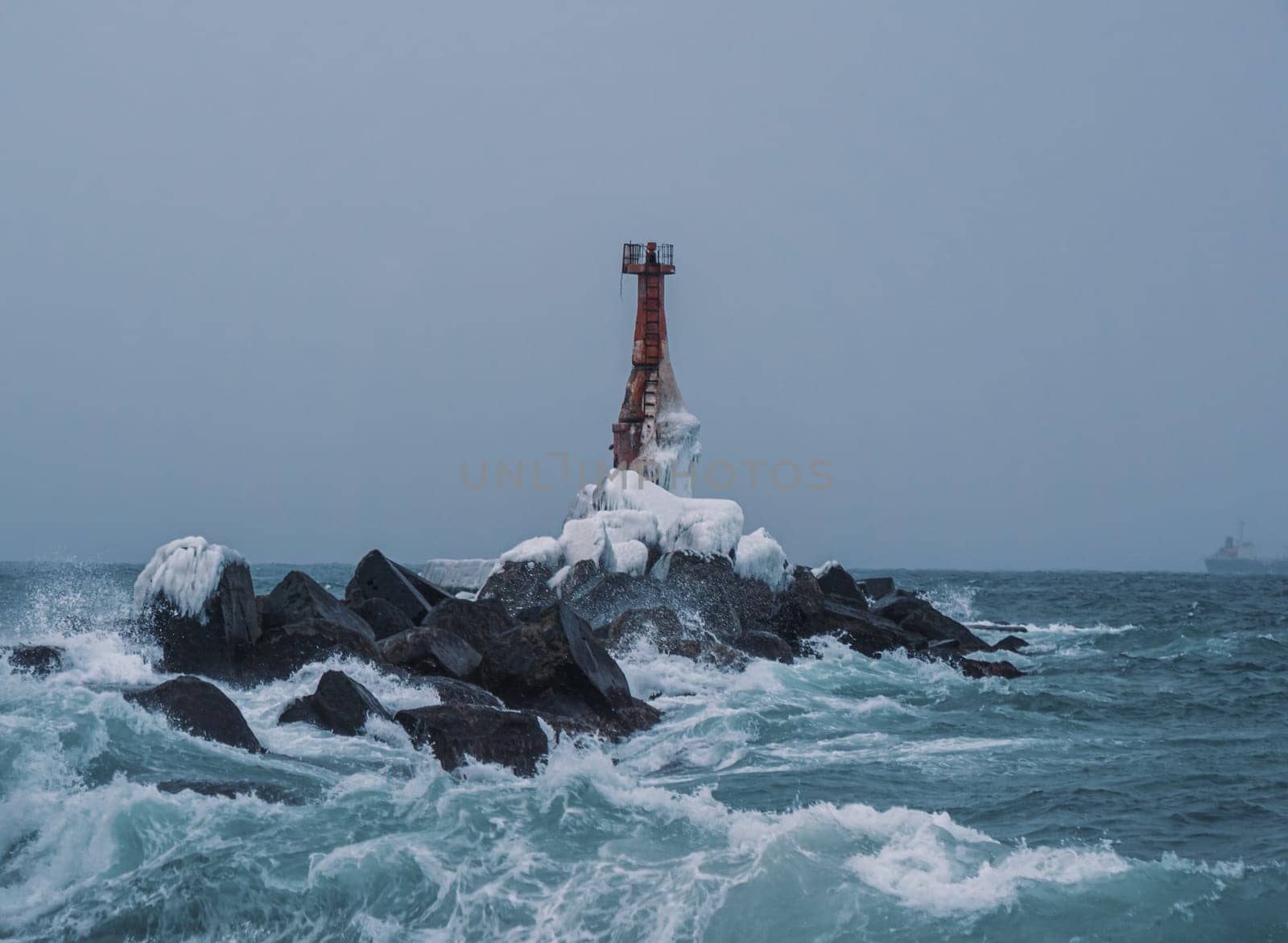 A solitary lighthouse stands firm on a rocky shore as waves crash against the rocks. The overcast sky and turbulent sea create a dramatic and moody atmosphere..