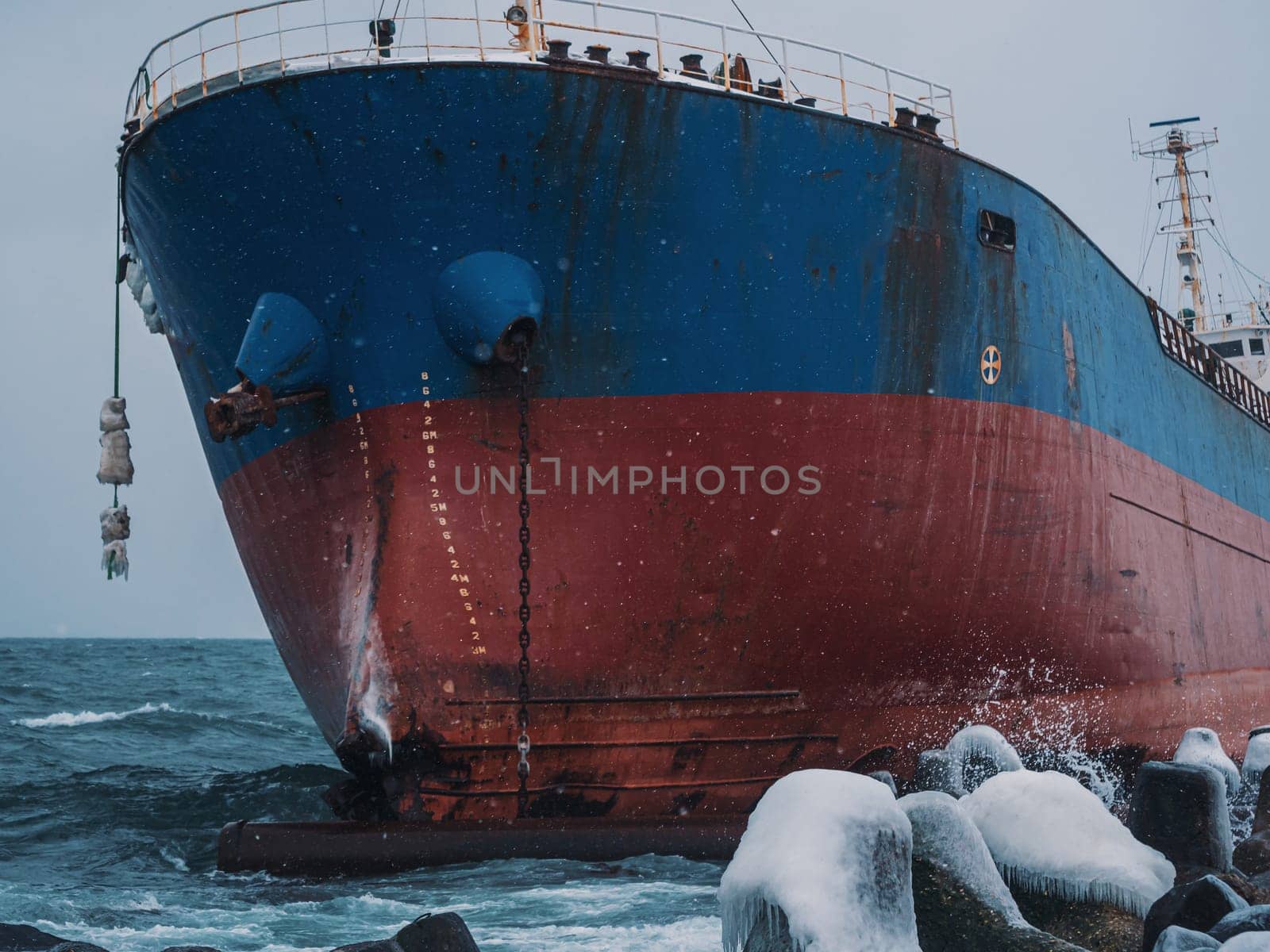 Cargo ship strands aground on a rocky shore after a storm, surrounded by snow and heavy waves.