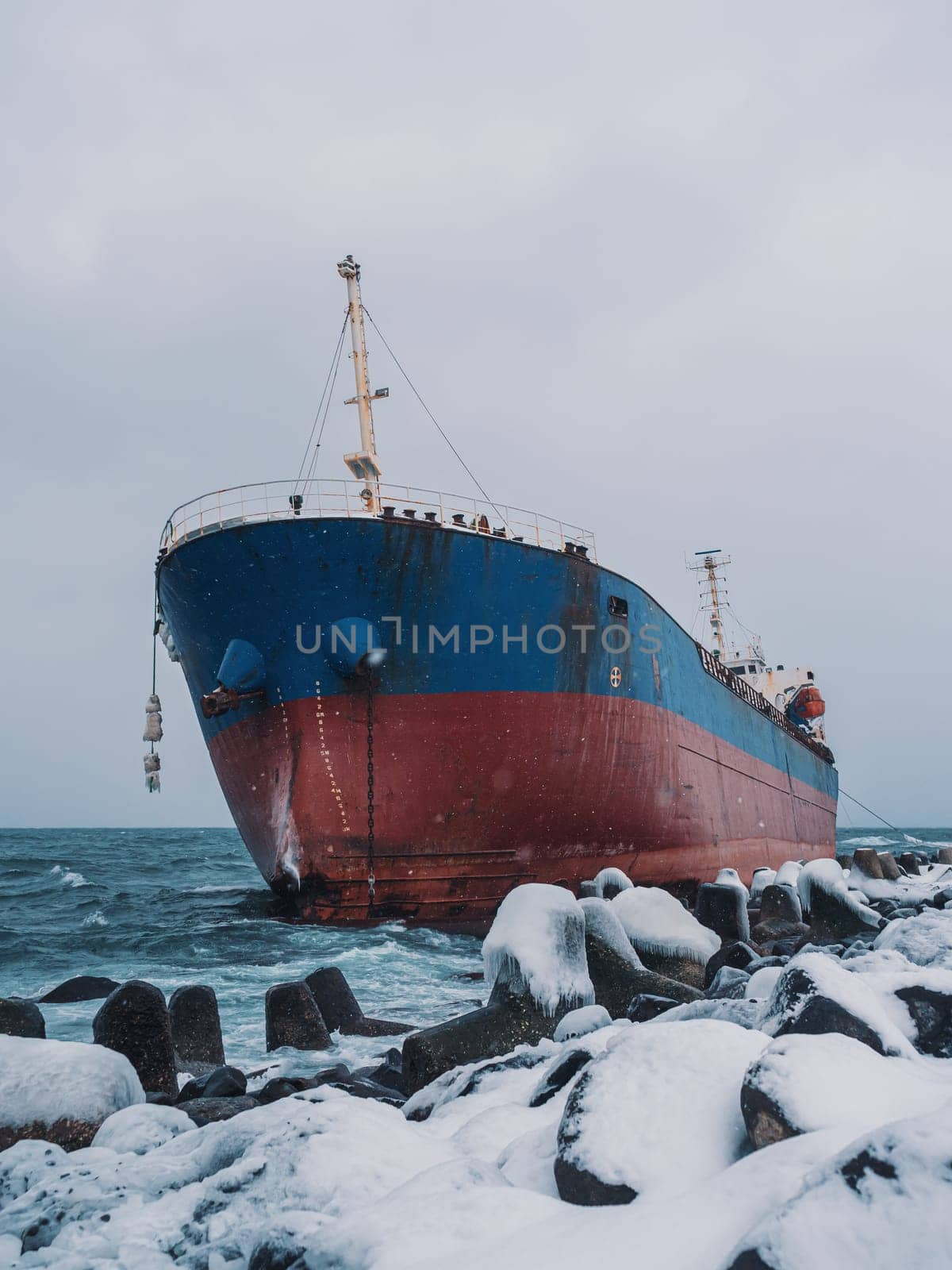 Cargo ship strands aground on a rocky shore after a storm, surrounded by snow and heavy waves.